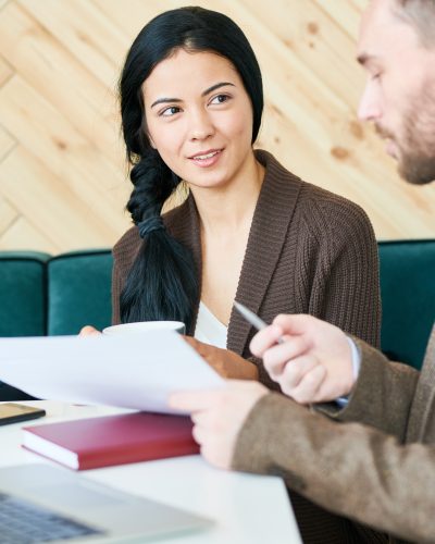 Portrait of pretty young businesswoman listening to her colleague, partner or client sitting at cafe table and discussing marketing strategy during meeting at coffee break